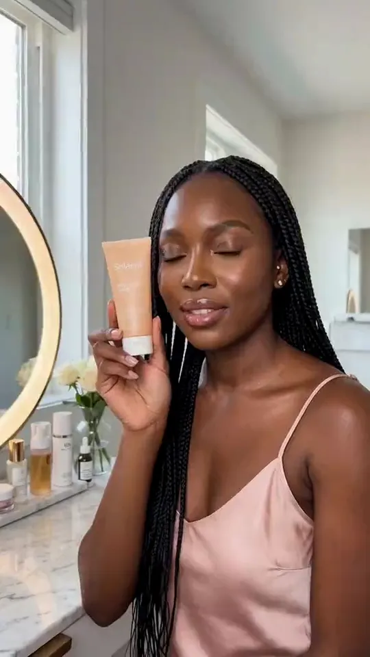 Clip from the One product, three AI influencers template: Vertical lifestyle video at her lit vanity: she holds the sunscreen tube up beside her cheek, turns the label toward the lens and says, warmly and clearly: "This is Solenne's daily glow SPF fifty. It goes on sheer, with no white cast." Then she smiles. Handheld phone camera, soft daylight. No music, no subtitles, no on-screen text.