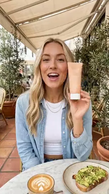 Clip from the One product, three AI influencers template: Vertical lifestyle video on the sunny café patio: she holds the sunscreen tube up beside her face, label toward the lens, and says, brightly and clearly: "Brunch on the patio means sunscreen. Solenne's SPF fifty is light and non-greasy." Then she laughs and sets it by her coffee. Handheld phone camera, warm sunlight. No music, no subtitles, no on-screen text.
